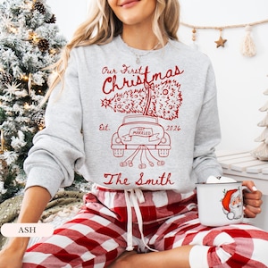 Pu&ograve; includere: Una felpa girocollo grigio chiaro con una stampa grafica rossa di un'auto che trasporta un albero di Natale. Il testo recita Our First Christmas, Est. 2026, Just Married e The Smith. Abbinata a pantaloni a quadri rossi e bianchi.