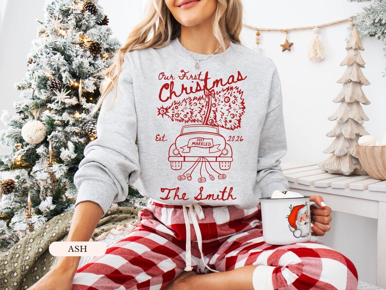 Pu&ograve; includere: Una felpa girocollo grigio chiaro con una stampa grafica rossa di un'auto che trasporta un albero di Natale. Il testo recita Our First Christmas, Est. 2026, Just Married e The Smith. Abbinata a pantaloni a quadri rossi e bianchi.