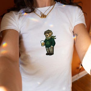 May include: White t-shirt featuring a cartoon bear wearing a green sweater, scarf, and cap, holding a mug of beer. The bear has a shamrock on its sweater. The design is centered on the shirt.