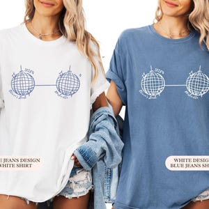 May include: Two t-shirts, one white and one blue, each with a graphic of two disco balls linked by a line. The text "Olivia's Bach Nashville" is printed below. The white shirt has a blue design, and the blue shirt has a white design. The shirts are casual wear.