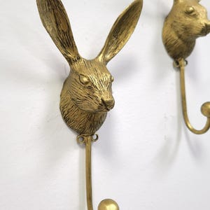 May include: Two golden-colored wall hooks shaped like rabbit heads. The hooks have long ears and detailed fur texture. Each hook has a curved, downward-facing arm ending in a small ball. The hooks are mounted on a white wall.