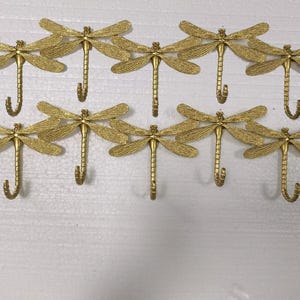 May include: A set of ten gold-colored dragonfly wall hooks. Each hook is designed with a dragonfly shape, featuring detailed wings and a curved hook for hanging items. The hooks are mounted on a white surface.