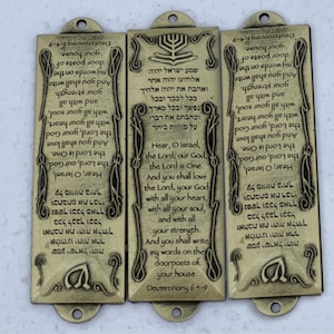 May include: Three gold-colored metal plaques with Hebrew text and English translations of the Shema prayer. The plaques are decorated with a menorah design and have a hole at the top for hanging.