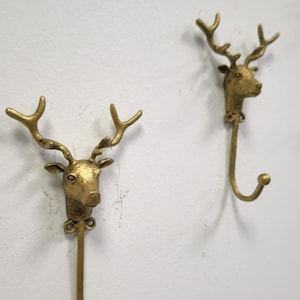 May include: Two gold-colored metal wall hooks, each featuring a deer head design. The hooks are mounted on a white wall. The deer heads have detailed antlers and are attached to a curved hook.