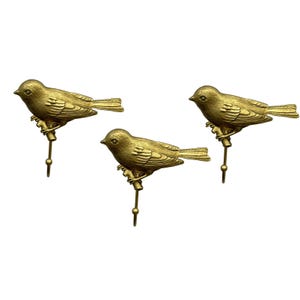 May include: A set of three metallic gold-toned wall hooks, each featuring a detailed bird perched on a small branch. The birds are depicted in profile with textured wings and tail feathers, and each hook terminates in a small, rounded ball tip.