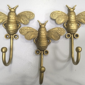May include: Three gold-colored bee-shaped wall hooks. Each hook features a detailed bee design with outstretched wings and a curved hook below. The hooks are made of metal and are suitable for hanging items. The hooks are approximately 5 inches in height.