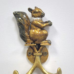 May include: A brass squirrel-shaped wall hook. The squirrel is in a sitting position with a fluffy tail and is mounted on a round base with two hooks. The squirrel is gold and brown with a white collar.
