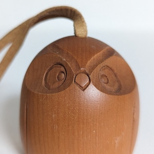 Può includere: Un ornamento di legno a forma di gufo con un cordino in pelle marrone. Il gufo ha un viso intagliato con occhi, un becco e due piccoli piedi.