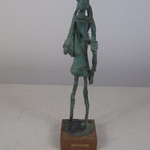 Puede incluir: Una escultura de bronce de una figura de pie con extremidades alargadas y una prenda drapeada. La figura está montada sobre una base de madera con una pequeña placa. La escultura tiene un aspecto envejecido, lo que sugiere que es antigua.