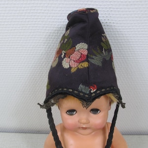 May include: A vintage doll wearing a dark blue bonnet with floral embroidery in pink, red, and green. The bonnet has a scalloped edge with black trim and black braided ties. The doll has blonde hair and a pale complexion.