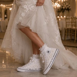 May include: A pair of white high-top sneakers featuring lace floral appliqués, satin ribbon laces, and a circular logo patch on the side. The sneakers are worn with white ruffled ankle socks, paired with a long white lace wedding dress.