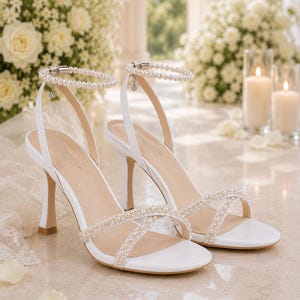 May include: Elegant white sandals with pearl accents. These heeled sandals feature a delicate ankle strap and crisscross straps across the foot, adorned with small pearls. The heels are approximately 3 inches tall, perfect for special occasions.