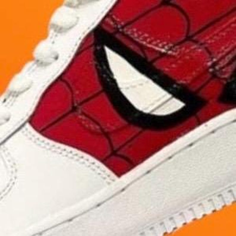 Puede incluir: Primer plano de una zapatilla blanca con un dise&ntilde;o de Spiderman rojo y negro. El dise&ntilde;o presenta un patr&oacute;n de telara&ntilde;a y el ojo de Spiderman.