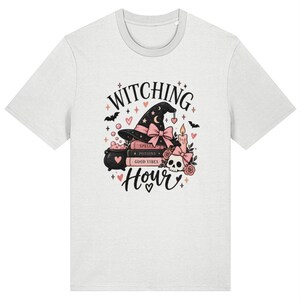 May include: A light gray t-shirt featuring a graphic print with the text "WITCHING Hour". The design includes a black witch hat with a pink bow, a stack of books labeled "SPELLS", "POTIONS", and "GOOD VIBES", a bubbling cauldron, a skull, and a candle.