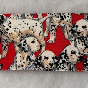 May include: A rectangular fabric item featuring a repeating pattern of Dalmatian dogs on a red background. The dogs are depicted in various poses, with their distinctive black spots and tan markings. The item appears to be a wallet or a similar accessory.