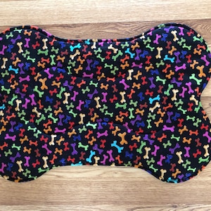 May include: Bone-shaped pet bed with a black background and a pattern of colorful bones in orange, yellow, purple, green, and blue. The bed is padded and has rounded edges.