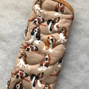 Puede incluir: Manopla de horno acolchada de color beige, adornada con un estampado repetido de perros Basset Hound. Los perros están representados en marrón, negro y blanco. La manopla tiene un ribete marrón y está diseñada para la cocina.