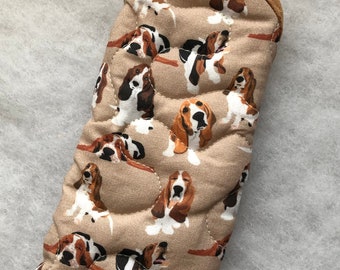 May include: Tan oven mitt featuring a repeating pattern of Basset Hound dogs in various poses. The mitt has a quilted design and a brown trim along the top edge. The dogs are in shades of brown, black, and white.