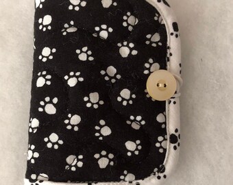 Può includere: Una pochette in tessuto nero e bianco con un motivo a forma di impronta di zampa. La pochette ha un bordo bianco e una chiusura con un unico bottone.