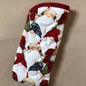 May include: A quilted oven mitt featuring a repeating pattern of Christmas gnomes with red hats and beards on a white background. The mitt has a red trim and is designed for kitchen use.