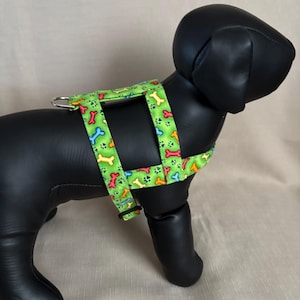 May include: A green dog harness with a colorful bone and paw print pattern. The harness is displayed on a black dog mannequin. The harness has a metal ring for leash attachment and adjustable straps.