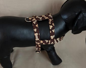 Könnte beinhalten: Ein braunes Hundegeschirr mit einem sich wiederholenden Muster aus weißen Hundeknochen. Das Geschirr befindet sich auf einer schwarzen Hunde-Mannequin. Es hat einen Metallring zur Befestigung der Leine.