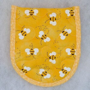 Puede incluir: Un agarrador de olla amarillo con un patr&oacute;n de abejas. El agarrador tiene un borde amarillo y est&aacute; acolchado con hilo blanco.