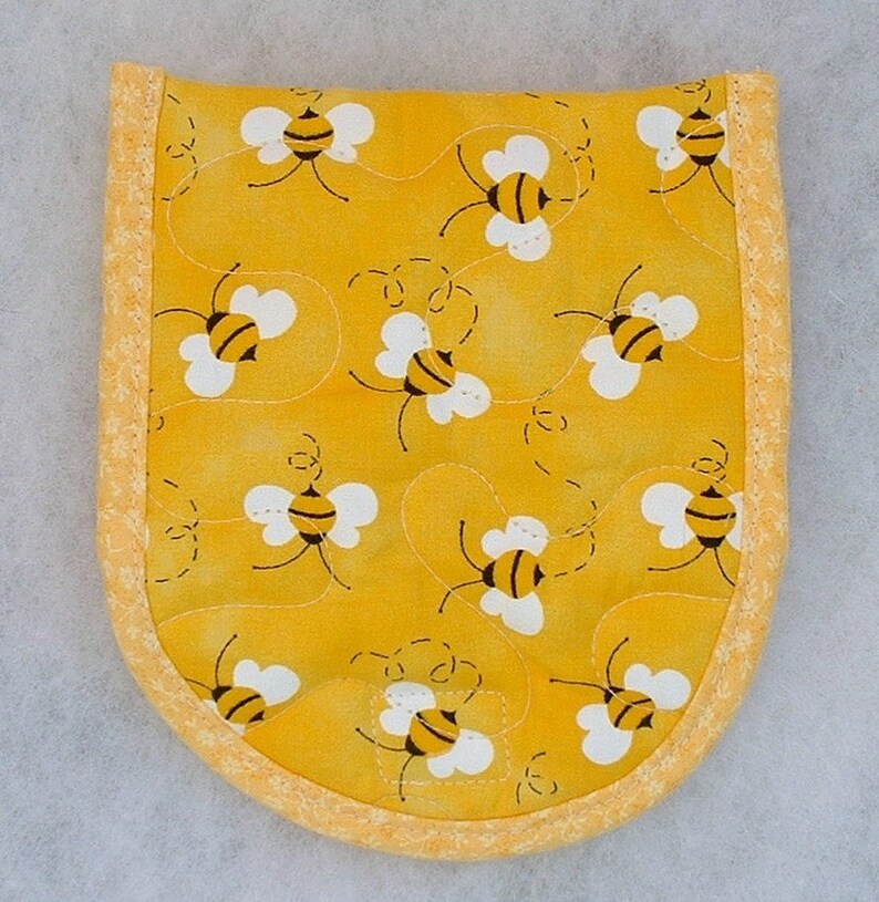 Puede incluir: Un agarrador de olla amarillo con un patr&oacute;n de abejas. El agarrador tiene un borde amarillo y est&aacute; acolchado con hilo blanco.