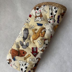 May include: A quilted fabric glasses case featuring a cream background with a repeating pattern of various dog breeds, white bones, red dog houses, and the word WOOF in yellow. The top edge is finished with a textured tan woven fabric trim.