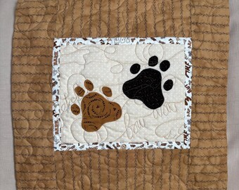 Könnte beinhalten: Ein braunes und hellbraunes gestepptes Kissen mit einem braunen und schwarzen Pfotenabdruck-Design. Das Kissen hat einen weißen Hintergrund mit braunen Punkten und den Text "Paw Prints" in einer kursivschrift.