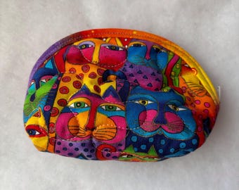 Puede incluir: Un estuche de tela semicircular con un patrón acolchado vibrante de caras de gatos estilizadas en colores llamativos como amarillo, azul, rosa y morado. El diseño incluye formas geométricas repetitivas y lunares en toda la superficie.
