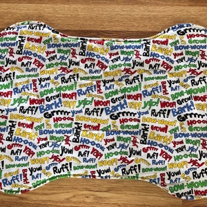 May include: White bone-shaped fabric placemat with a repeating pattern of dog-related words in black, red, yellow, and green. The words include "Bark", "Woof", "Growl", "Yip", "Ruff", and "Bow-Wow".
