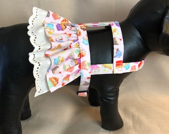 Könnte beinhalten: Ein weißes Hundegeschirr mit Cupcake-Druck und einer weißen Spitzenrüsche. Das Geschirr hat verstellbare Träger und einen Schnallenverschluss.
