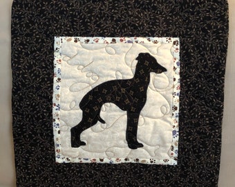 Könnte beinhalten: Ein schwarzes und weißes gestepptes Kissen mit einer schwarzen Silhouette eines Hundes und dem Wort "Hund" in Kursivschrift. Das Kissen hat einen schwarzen Hintergrund mit einem weißen Rand und ein weißes Innenquadrat mit einem braunen, weißen und blauen Rand. Das Innenquadrat hat ein Muster aus Pfotenabdrücken.