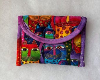Könnte beinhalten: Eine kleine, rechteckige Stofftasche mit einem lebendigen, bunten Muster aus stilisierten Katzengesichtern in den Farben Rot, Blau, Grün, Orange und Violett. Die Tasche hat einen geschwungenen Klappenverschluss mit einem violetten Stoffrand.