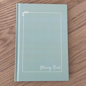 Op de afbeelding: Een lichtgroen herinneringsboek met een witte rand en de woorden "Memory Book" in cursief. De omslag heeft een eenvoudig bloemmotief in de linkerbovenhoek. Het boek is gesloten en ligt op een houten oppervlak.