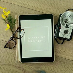 Op de afbeelding: Een iPad met de tekst "A Year of Memories" op het scherm, een vintage camera, een bril en een kleine vaas met gele bloemen zijn gerangschikt op een houten oppervlak. De iPad zit in een beschermhoes.