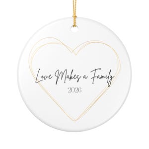 Könnte beinhalten: Weißer Keramik-Weihnachtsschmuck mit goldfarbener Herzumrandung und goldenem Aufhängeband. Der Text "Love Makes a Family" und das Jahr "2026" sind in schwarzer Schrift innerhalb des Herzens geschrieben.