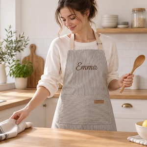 May include: A grey and white pinstriped apron featuring a large front pocket with a small rectangular leather-like patch and the name Emma embroidered in cursive script on the chest. The apron has light-colored neck straps and waist ties.