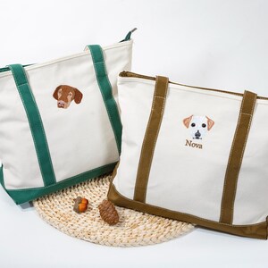 May include: Two canvas tote bags with embroidered dog portraits. One bag has green handles and trim, the other has brown. The bag on the right has the name "Nova" embroidered below the dog portrait. Both bags are white with a woven base.