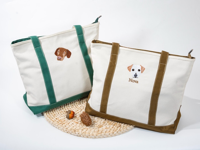 May include: Two canvas tote bags with embroidered dog portraits. One bag has green handles and trim, the other has brown. The bag on the right has the name "Nova" embroidered below the dog portrait. Both bags are white with a woven base.