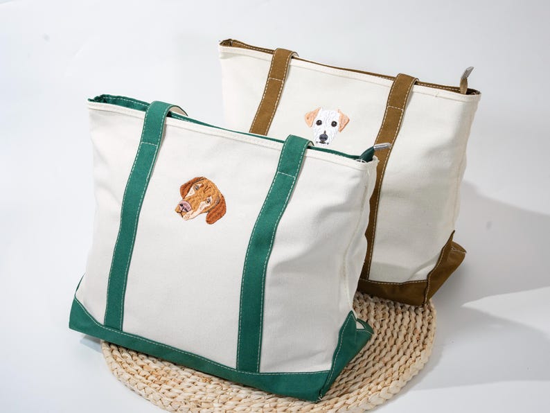 May include: Two canvas tote bags with embroidered dog portraits. One bag has green accents and the other has brown accents. Both bags are white with a zipper closure and sturdy handles. The bags are sitting on a woven mat.