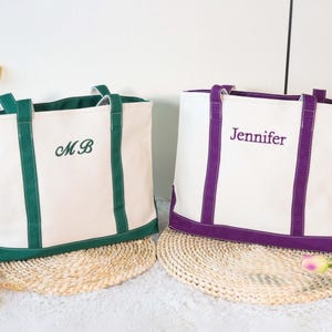 Puede incluir: Dos bolsas de mano de lona blanca con ribetes y asas en contraste. Una bolsa tiene ribetes verdes y las iniciales "MB" bordadas. La otra bolsa tiene ribetes morados y el nombre "Jennifer" bordado. Ambas bolsas están sobre una estera tejida.