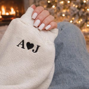 Op de afbeelding: Crèmekleurige sweatshirt met geborduurde initialen "A" en "J" met een hartsymbool. De persoon draagt een blauwe spijkerbroek. De achtergrond omvat een open haard en een kerstboom met lichtjes.