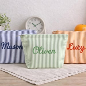 Puede incluir: Tres neceseres a rayas en azul, verde y naranja, cada uno bordado con un nombre: Mason, Oliver y Lucy. Las bolsas están sobre un paño de lino, con un reloj y una planta en el fondo.