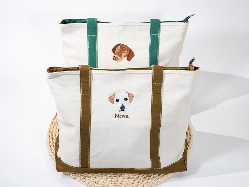 May include: Two canvas tote bags with embroidered dog portraits. The top bag has a green handle and a brown dog portrait. The bottom bag has brown handles and a white dog portrait with the name "Nova" embroidered below.