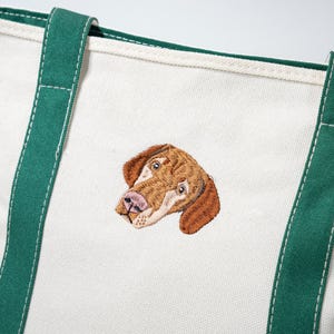 May include: Off-white canvas tote bag with green trim and handles. The bag features an embroidered image of a dog's head in shades of brown, tan, and pink. The handles are a deep green color.