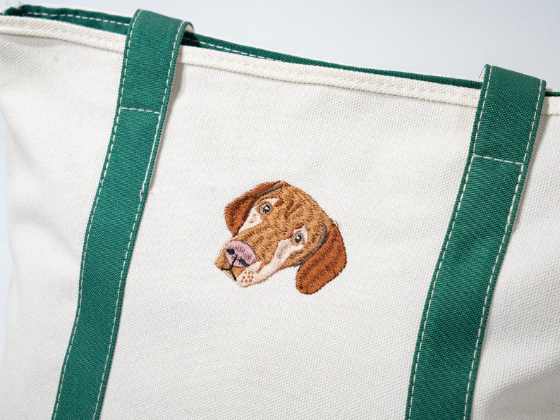 May include: Off-white canvas tote bag with green trim and handles. The bag features an embroidered image of a dog's head in shades of brown, tan, and pink. The handles are a deep green color.