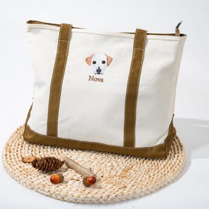 May include: Cream-colored tote bag with brown accents and handles. The bag features an embroidered dog face with the name "Nova" below. The bag is sitting on a woven mat with decorative acorns and a pinecone.