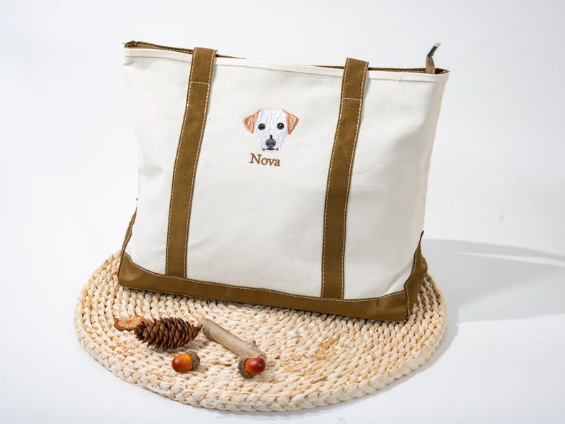 May include: Cream-colored tote bag with brown accents and handles. The bag features an embroidered dog face with the name "Nova" below. The bag is sitting on a woven mat with decorative acorns and a pinecone.
